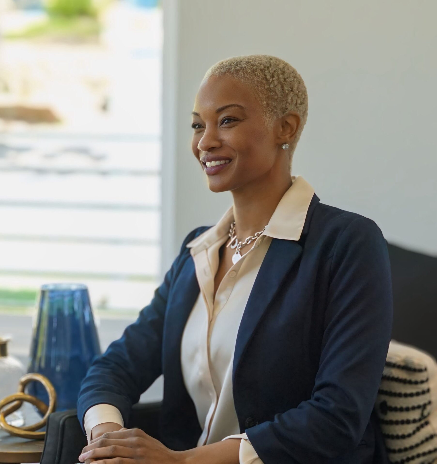A professional woman with short blonde hair, wearing a navy blazer and cream blouse, smiling confidently while seated at a table with decorative elements in the background. The setting is modern and well-lit, evoking a calm, professional atmosphere.